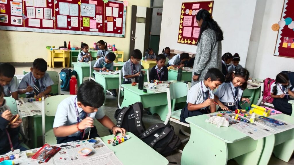 A classroom at Vicon School, the best school in Raipur with extracurricular activities, with young students engaged in art and craft activities. The children are sitting at individual desks, painting and creating crafts with various materials. A teacher is assisting the students, and the classroom walls are decorated with colorful posters and student work. The students are wearing school uniforms and appear focused and happy.